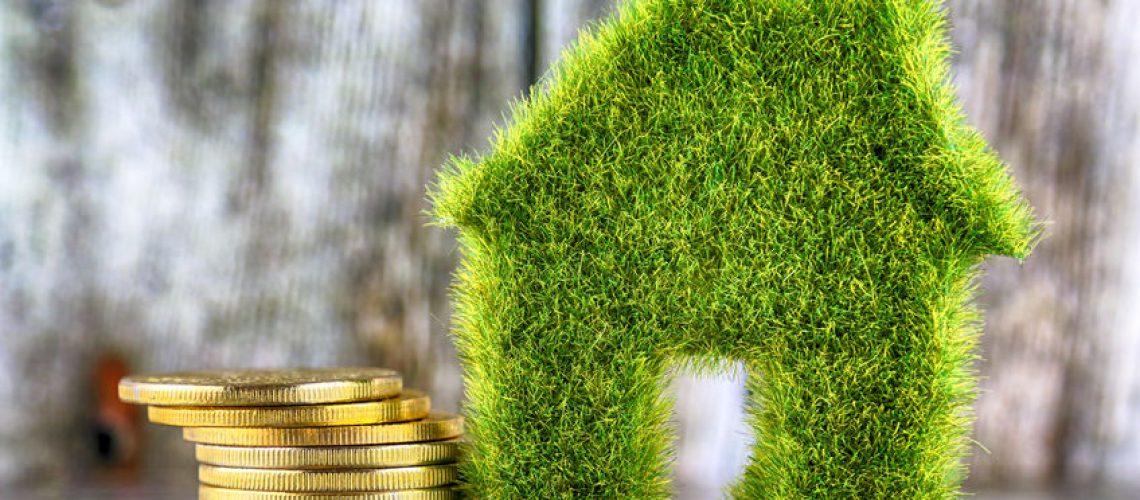 Miniature green eco house icon with grass and golden coins on wooden background. Renewable energy concept. Energy saving in the household.