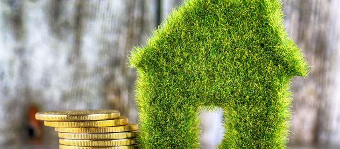 Miniature green eco house icon with grass and golden coins on wooden background. Renewable energy concept. Energy saving in the household.