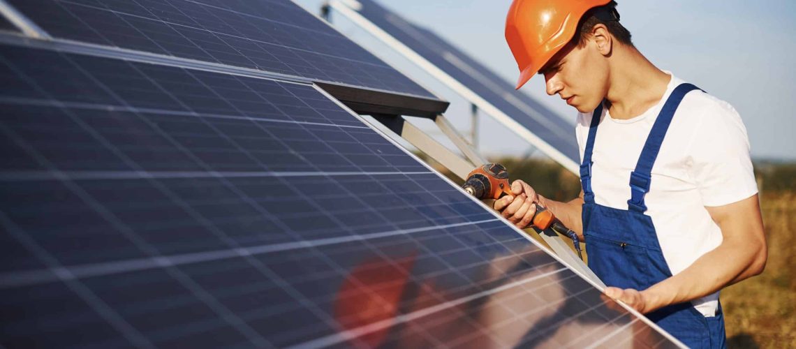 Using cordless screwdriver. Male worker in blue uniform outdoors with solar batteries at sunny day.