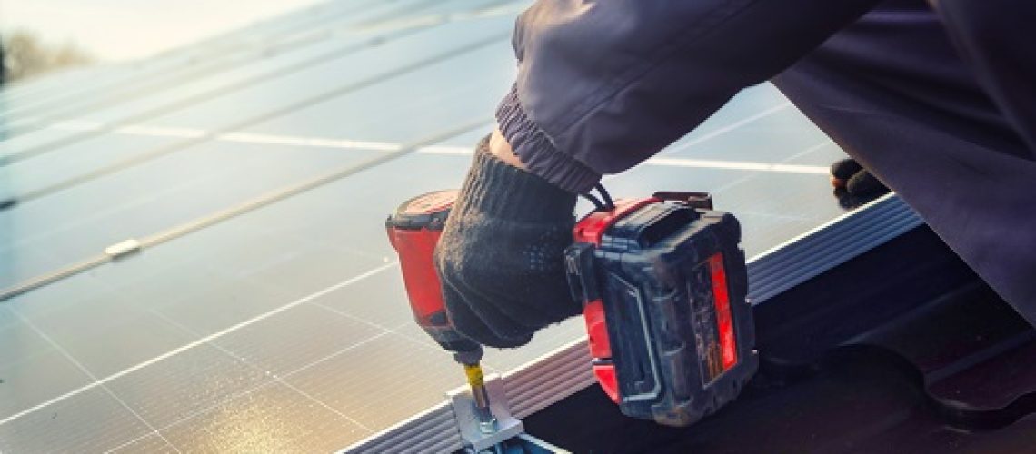 Solar panel technician with drill installing solar panels on roof.  technician in blue suit installing photovoltaic blue solar modules with screw. Electrician panel sun sustainable resources