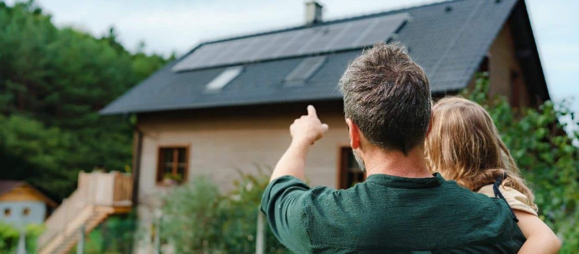 Une homme portant sa fille dans son jardin montrant leur maison qui a été construite grâce aux aides à la rénovation énergétique en 2025
